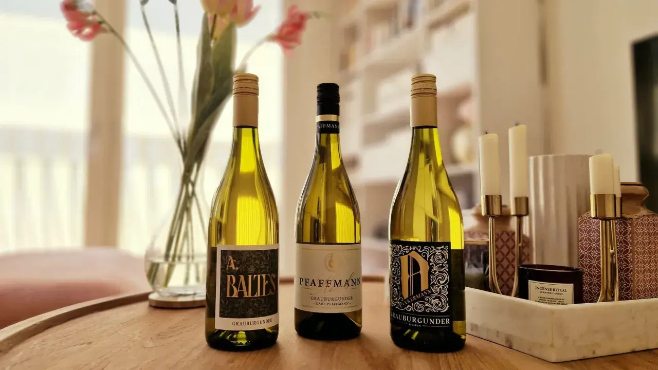 Drei Flaschen Grauburgunder Wein stehen auf einem Holztisch, daneben Blumen und Kerzen.