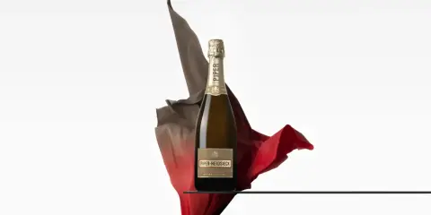 Eine Flasche Piper Heidsieck Cuvée Sublime, umhüllt von fließenden roten und braunen Stoffen, auf einer Glasplatte.