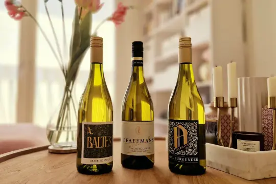 Drei Flaschen Grauburgunder Wein stehen auf einem Holztisch, daneben Blumen und Kerzen.