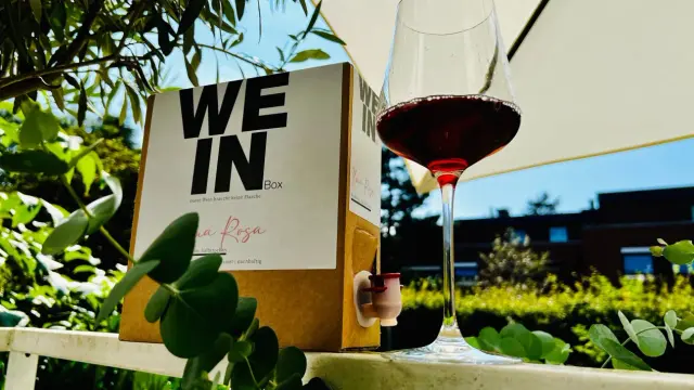 Ein Glas Rotwein steht neben einem Karton mit der Aufschrift "WE IN Box". Der Wein im Karton ist eine praktische Alternative zur Flasche.