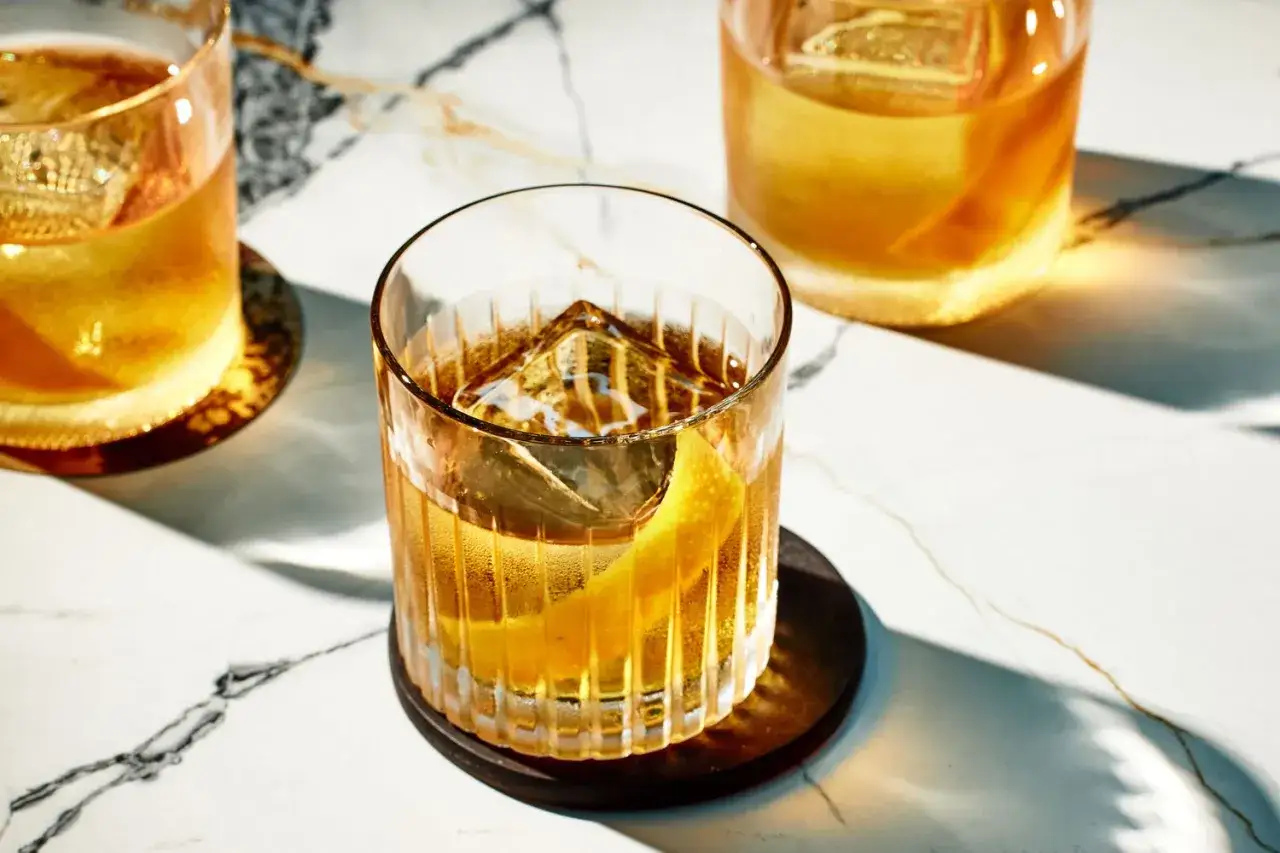 Drei klassische Whiskey-Cocktails mit großen Eiswürfeln und Zitronenzesten auf Marmortisch.