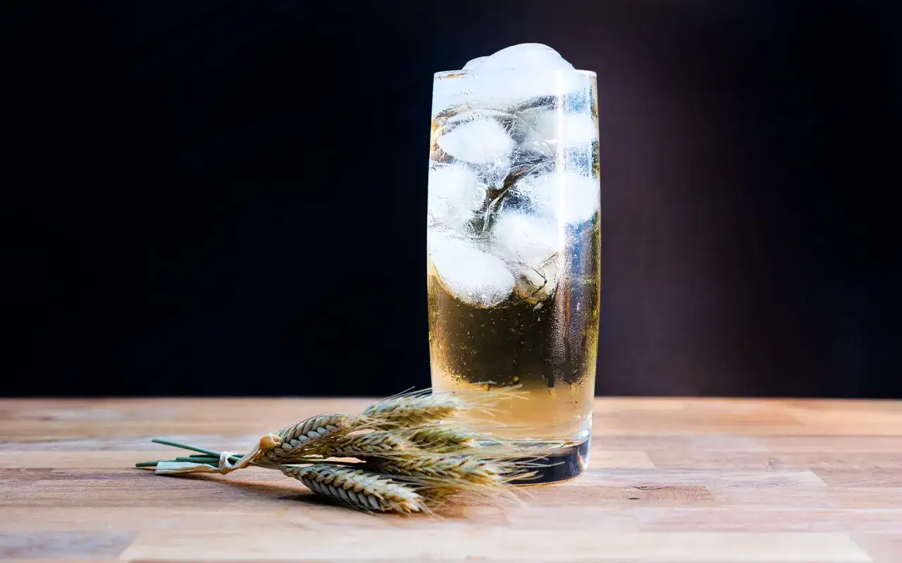 Ein erfrischender Whisky Soda mit Eiswürfeln, daneben ein Bündel Weizenähren auf Holztisch.