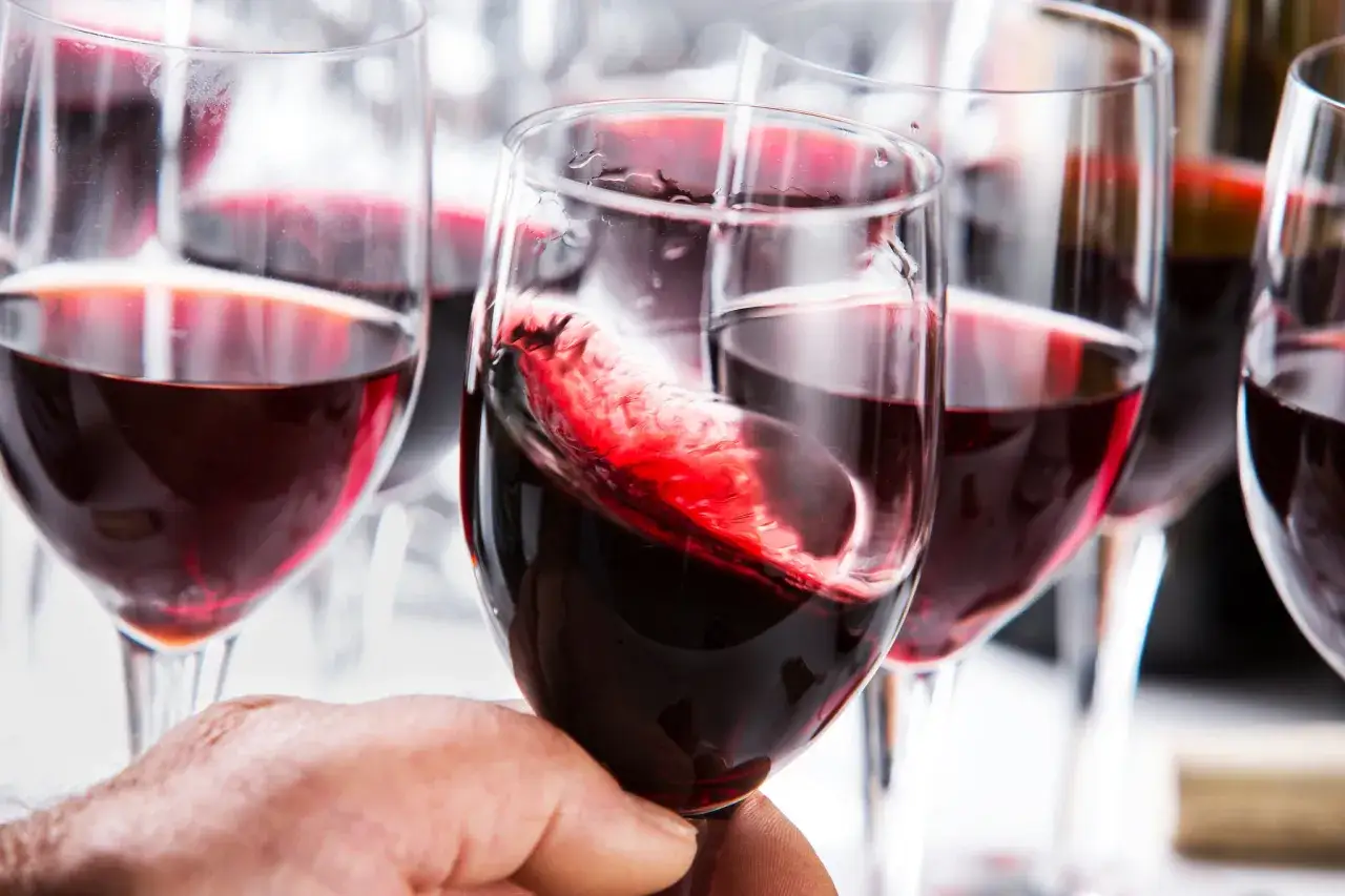 Ein Glas mit wirbelndem Grenache Rotwein wird von einer Hand gehalten. Mehrere Gläser mit tiefrotem Wein sind im Hintergrund zu sehen.