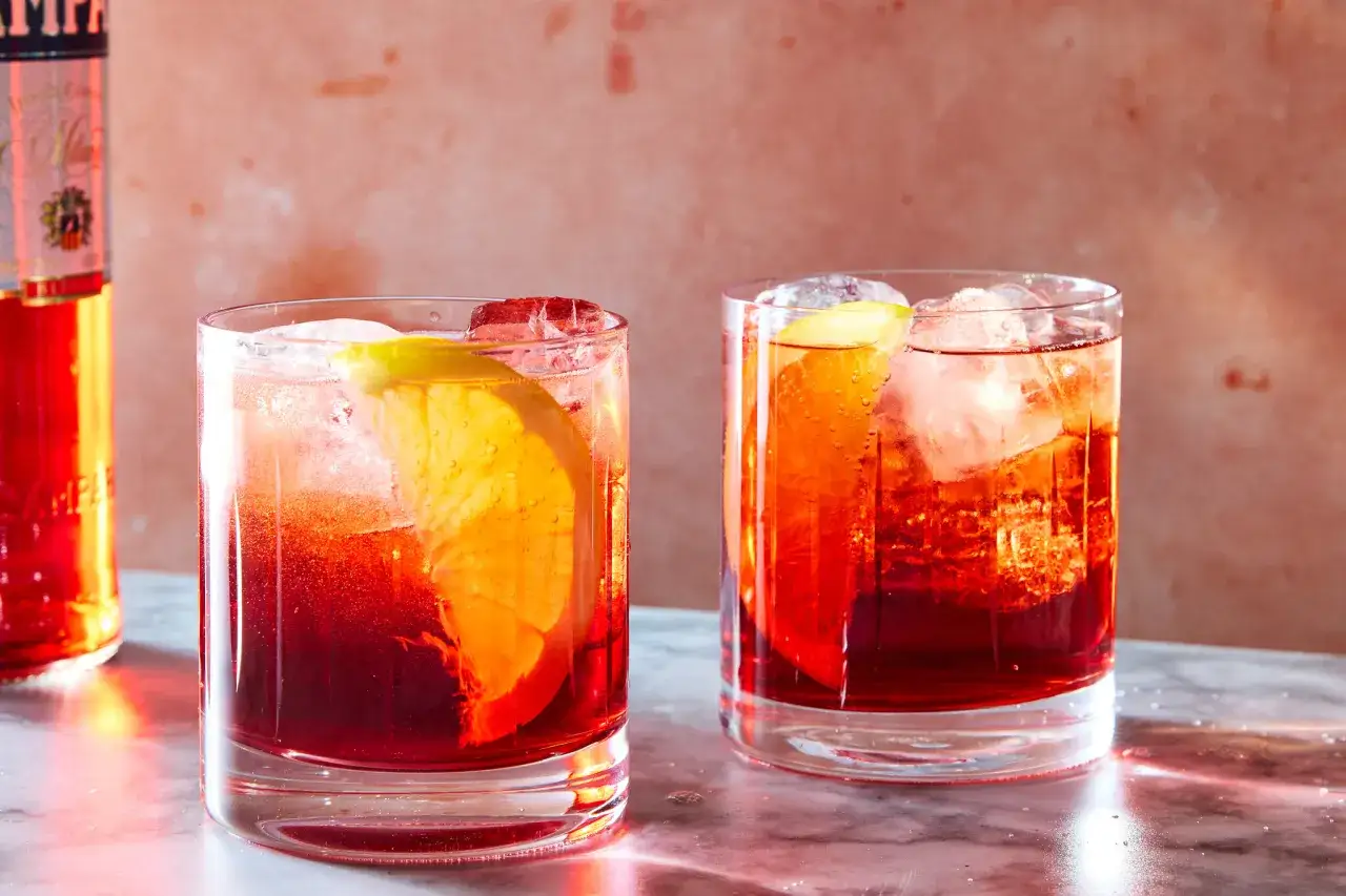 Zwei erfrischende Negroni Sbagliato Cocktails mit Eis und Orangenscheiben, daneben eine Flasche Campari.