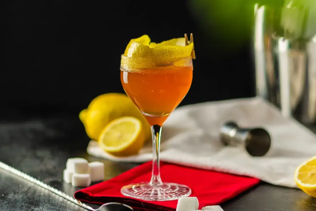 Elegantes Cognac-Cocktail mit Zitronenzeste, garniert mit einem kleinen Wäscheklammer-Detail. Zitronen und Zuckerwürfel deuten auf die Zubereitung hin.