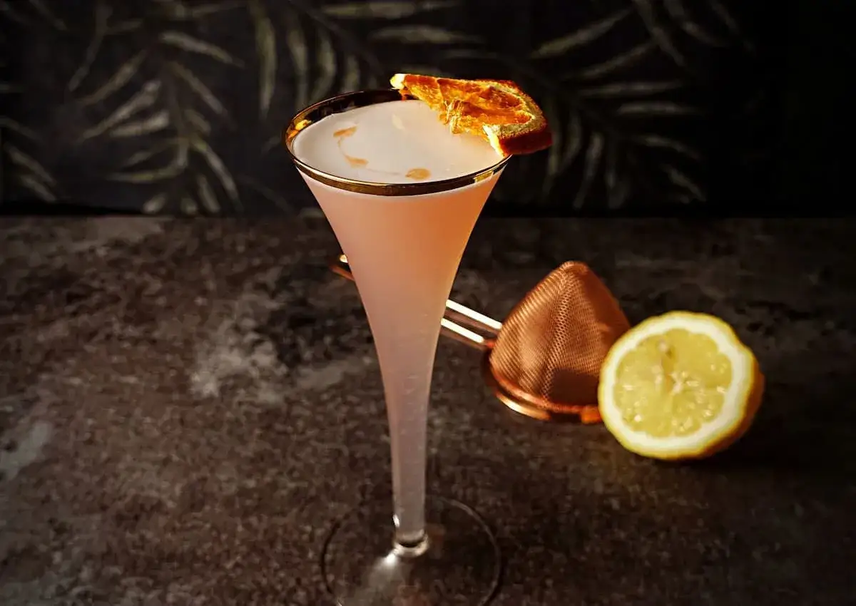 Ein rosafarbener Gin Sour Cocktail in einem eleganten Glas mit getrockneter Orangenscheibe, Zitrone und Kupfer-Barsieb.